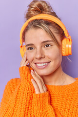 Wall Mural - Vertical shot of pleased young European woman focused into distance has cheerful expression dressed in knitted sweater via stereo headphones enjoys favorite music isolated over purple background