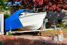 Recreational Boat On Trailer Stored On Driveway Of Residential Home. The Cruiser Is Covered With A Blue Tarp Tent For Protection.