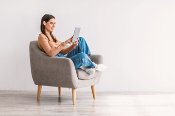 Happy young lady using tablet, studying or working remotely, having online conference, sitting in armchair, copy space