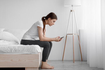 Canvas Print - Sad caucasian young woman suffering from depression and sitting in bed at home and reading message on smartphone
