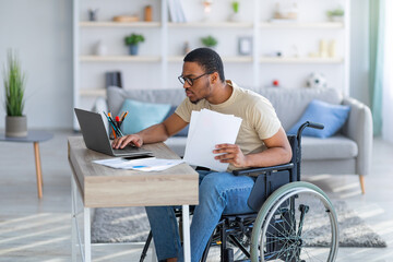 Wall Mural - Paraplegic black guy in wheelchair with documents using laptop to work online from home