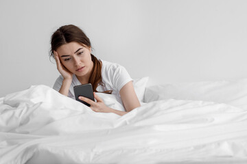 Wall Mural - Sad european young woman suffering from depression and read message on smartphone sit on white bed at home