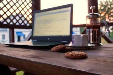 laptop with code on the screen, cup of coffee, cookies, french press, lies on the table in the gazeb