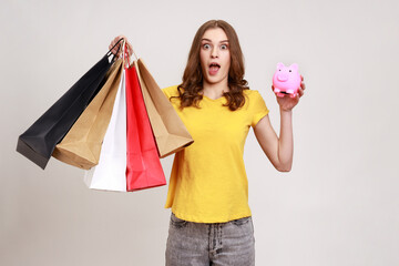 Poster - Portrait of astonished female with open mouth in yellow T-shirt holding shopping bags and piggy bank, save big mount of money, great purchase. Indoor studio shot isolated on gray background.