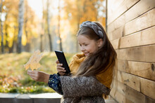 Smiling Girl Photographing Autumn Leaf Through Mobile Phone In Front Of Wooden Wall