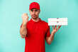Young delivery man taking pizzas isolated on blue bakcground showing fist to camera, aggressive facial expression.