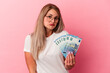 © Asier - Young russian woman holding bills isolated on pink background shrugs shoulders and open eyes confused.