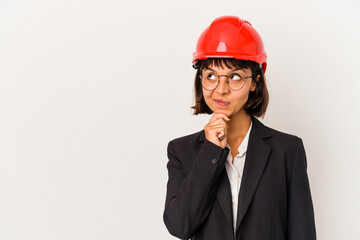 Wall Mural - Young architect woman with red helmet isolated on white background looking sideways with doubtful and skeptical expression.