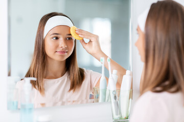 Canvas Print - beauty, hygiene and people concept - teenage girl with cleansing sponge cleaning facial skin and looking in mirror at bathroom