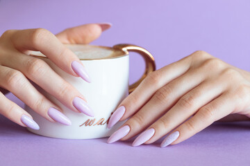 Wall Mural - Girl`s hand with a light purple manicure and a cup of cappuccino on a purple background, blank space