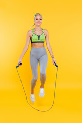 Wall Mural - full length view of happy woman in grey leggings and green sports bra jumping with skipping rope on yellow