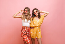 Photo Of Two Young Caucasian Women In Bright Clothes With Polka Dots Standing On Pink Background. Blonde And Brunette In Sunglasses Are Smiling Broadly With Their Teeth And Showing Sign Of Peace.