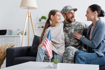 Canvas Print - Happy family hugging man in camouflage uniform on couch near american flag at home