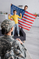 Canvas Print - Woman in military uniform with backpack near blurred family holding american flag in kitchen