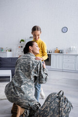 Canvas Print - Smiling woman hugging child near military backpack at home