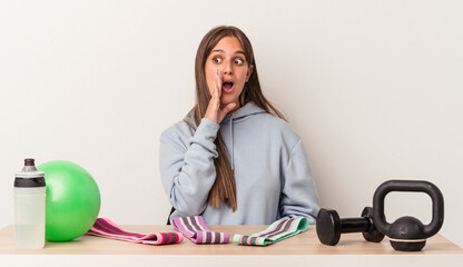 Wall Mural - Young caucasian woman sitting at a table with sport equipment isolated on white background is saying a secret hot braking news and looking aside