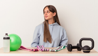 Wall Mural - Young caucasian woman sitting at a table with sport equipment isolated on white background gazing left, sideways pose.