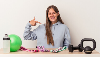 Wall Mural - Young caucasian woman sitting at a table with sport equipment isolated on white background person pointing by hand to a shirt copy space, proud and confident