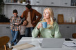 thoughtful woman with cup of tea sitting near laptop, documents, and blurred husband with kids in kitchen