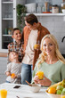 © LIGHTFIELD STUDIOS - cheerful woman looking at camera near blurred husband and sons in kitchen
