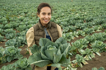 Portrait of smiling pretty young biracial horticulturist in vest standing with fresh big cabbage on field