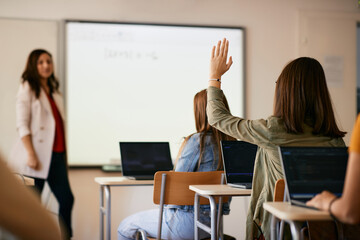 Rear view of student raising hand to ask a question during IT class at high school.