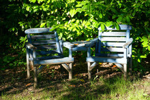 London, England - July 3 2021 - Two Rustic Blue Wooden Chairs In Dappled Sun Under A Large Green Tree In A Grassy Field.  Image Has Copy Space.