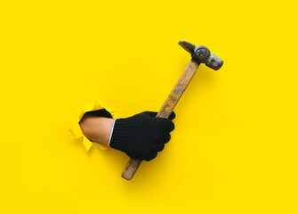 Wall Mural - A hand in a black knitted glove holds a clam hammer. Torn hole in yellow paper. The concept of a worker, labor migrant, a master of his craft. Copy space.