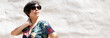 Portrait of a stylish Asian woman in beautiful beach dress with UV block sunglasses standing in front of a white plaster texture concrete wall under the sun. Vacation, Holidays, Santorini, Copy space.