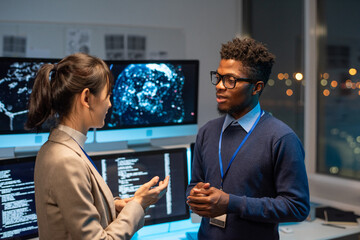 Poster - Two young multiracial colleagues having discussion against workplace with computer monitors in large openspace office