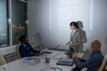 Wall Mural - Young confident Asian businesswoman with papers interacting with African male colleague sitting by desk in front of her