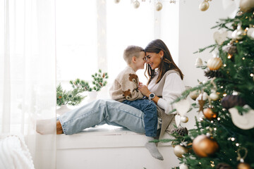 Happy mother with son sit on windowsill near Christmas tree