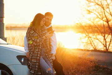 Wall Mural - Couple traveler stands by car at sunset in countryside. Girl uses mobile app map in smartphone to find out the way. Tourists checking location in smartphone map. Soft focus