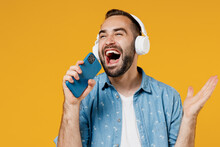 Young Expressive Happy Caucasian Man 20s In Blue Shirt White T-shirt Headphones Listen To Music Sing Song Record Voice On Mobile Phone Dictaphone Isolated On Plain Yellow Background Studio Portrait