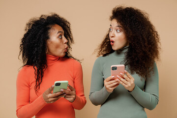 Wall Mural - Two shocked surprised young curly black women friends 20s wearing casual shirts clothes hold in hand use mobile cell phone look at each other isolated on plain pastel beige background studio portrait.