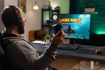 Wall Mural - Player winning video games with controller and headset in front of monitor. Man using joystick and headphones, playing online games on computer. Person celebrating game win for leisure.