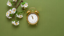 Decorative Flowering Cherry Branch And Golden Alarm Clock On Olive Green Paper Background. Spring Time. Top View, Flat Lay. Creative Layout.