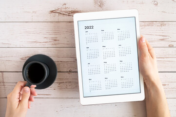 tablet computer with an open app of calendar for 2022 year and cup of tea or coffee in womans hands on wooden background. concept business or to do list goals with technology using. top view, flat lay