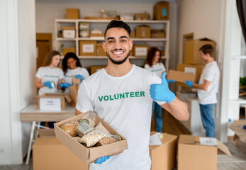 Wall Mural - Happy arab man at volunteer center holding donations paper box, showing thumb up and smiling at camera