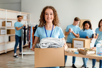 Wall Mural - Volunteering concept. Happy young woman in uniform holding cardboard box with clothes for donation