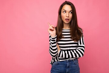 Wall Mural - Photo of young european positive thinking and dreaming beautiful brunette woman with sincere emotions wearing casual striped pullover and having an idea isolated on pink background with copy space for