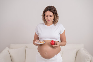 A pregnant woman is holding a red apple and donut. Choice of food.