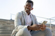 © ADDICTIVE STOCK - Smiling black businessman using smartphone on concrete steps