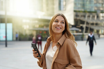 Wall Mural - Casual cheerful girl walking and holding smart phone in city street wearing a coat in spring time. City lifestyle people technology.