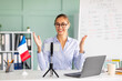 © Prostock-studio - Cheerful female tutor teaching French online using smartphone and laptop, talking to cellphone webcamera