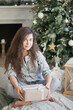 © Dina - Beautiful woman at home relaxed with a gift box sitting by the decorated christmas tree near fireplace. Christmas moments at cozy home decorated for holiday season.