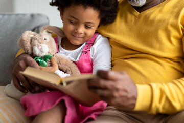 Wall Mural - Smiling small african american girl with toy reads book with elderly grandfather on sofa at home interior, cropped