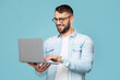 © Prostock-studio - Smiling attractive adult european male in glasses typing on computer, works remotely, isolated on blue background