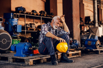 Wall Mural - An old, tired bearded factory worker in overalls is sitting on the pallet and taking a break from hard work. He is wiping sweat from his forehead.