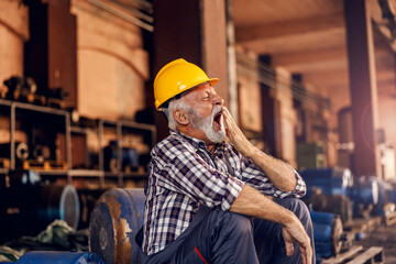 a tired senior worker is sitting next to the machines and yawning. after hard work, he must get some